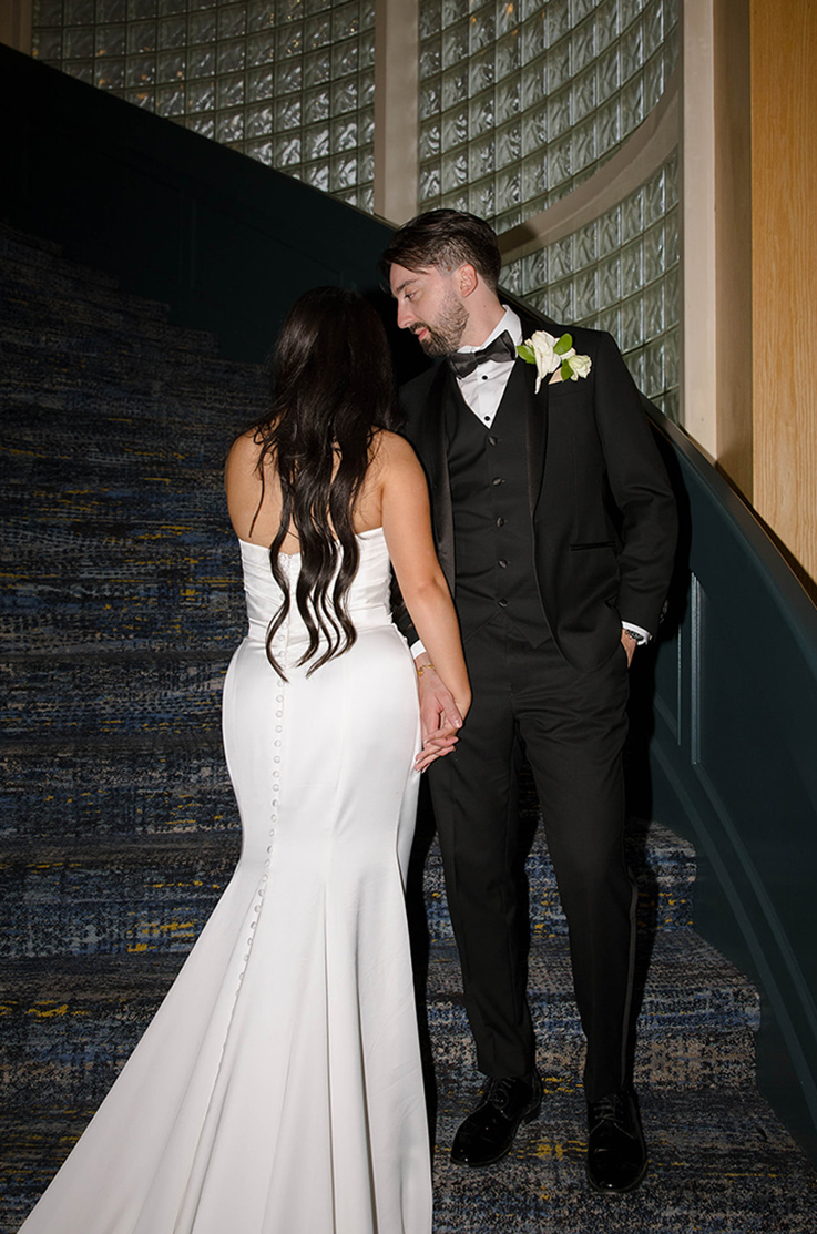 Back of bride in a draped fit and flare satin gown with buttons, holds hands with groom in a tux on spiral stairs indoors.