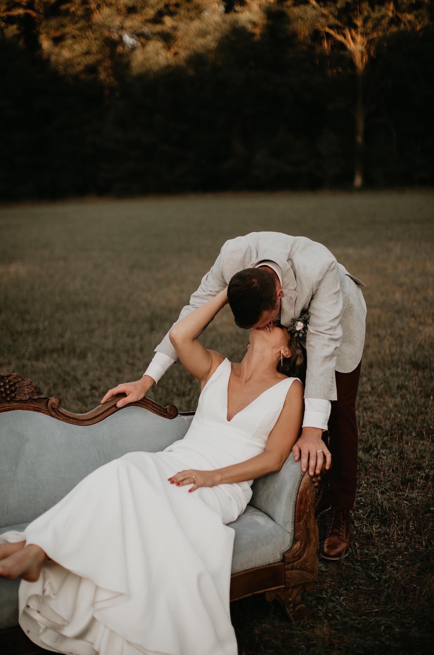 Bride sits back on a vintage couch outdoors in a V-neck crepe wedding gown kissing groom in an open field.