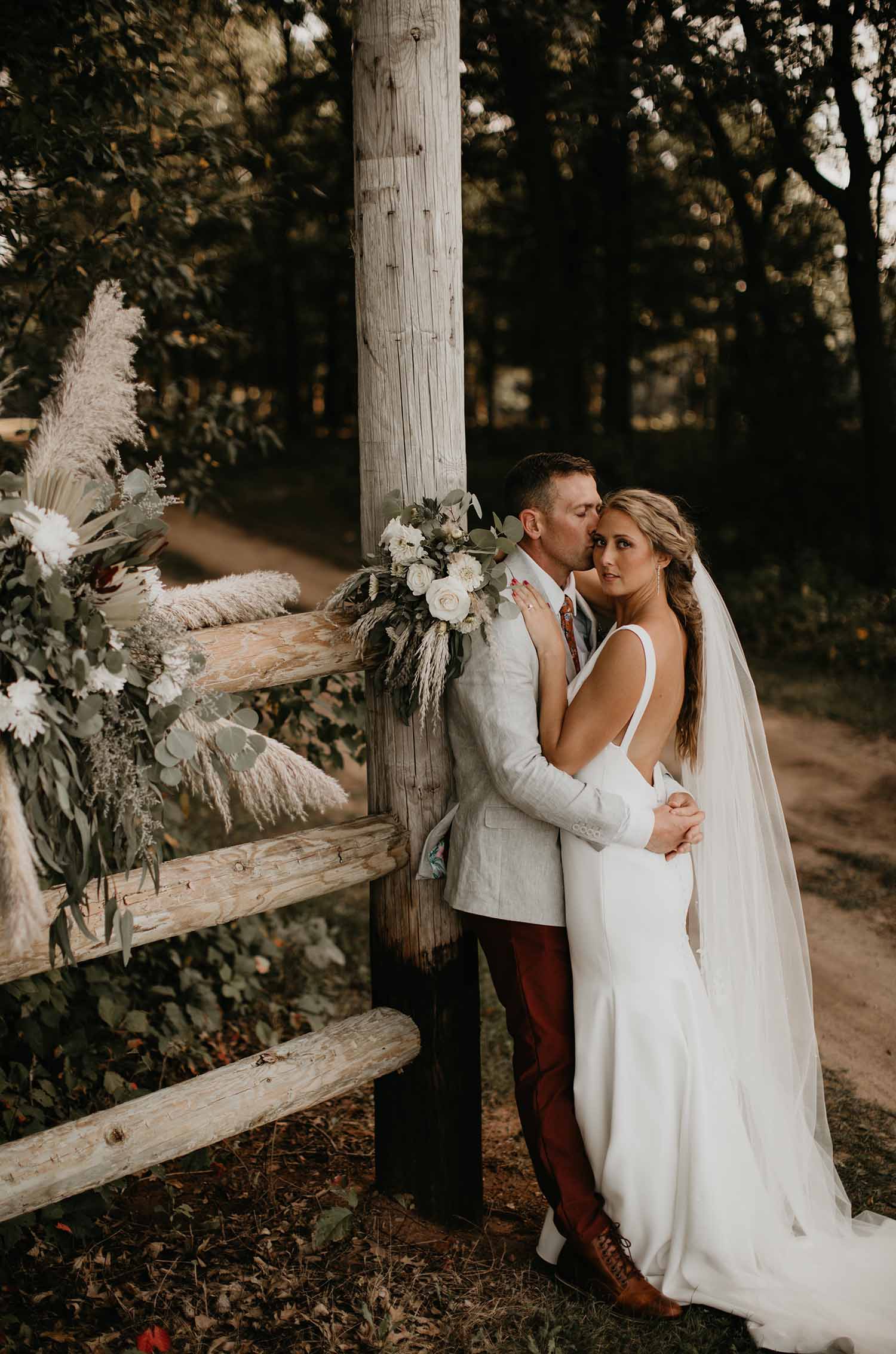Groom embraces bride in an open back crepe wedding gown with a bow at the back and a veil, against a decorated log fence. 