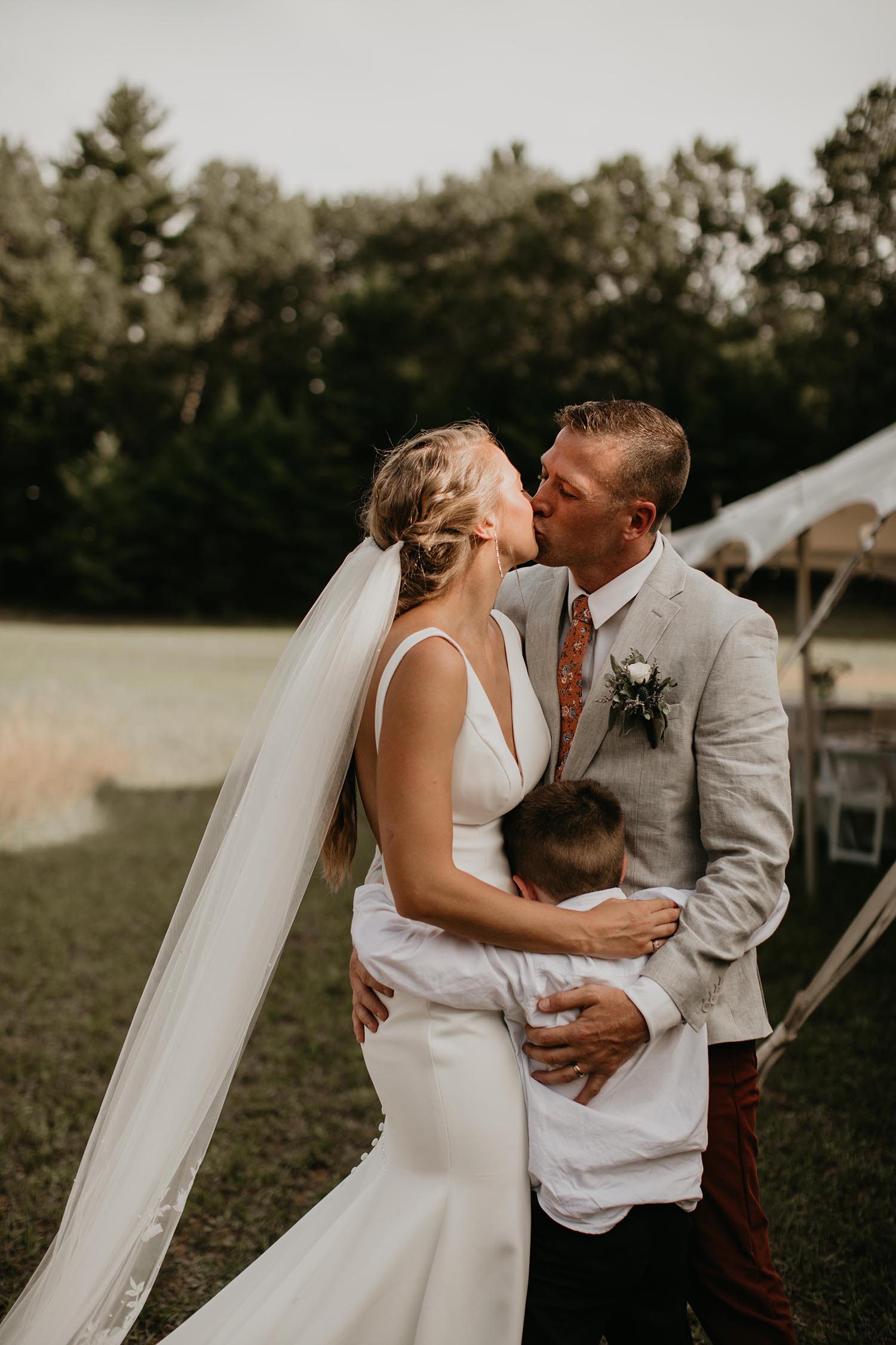Bride in a V-neck fit and flare gown and a veil, kisses groom in gray by an outdoor tent in a meadow setting. 