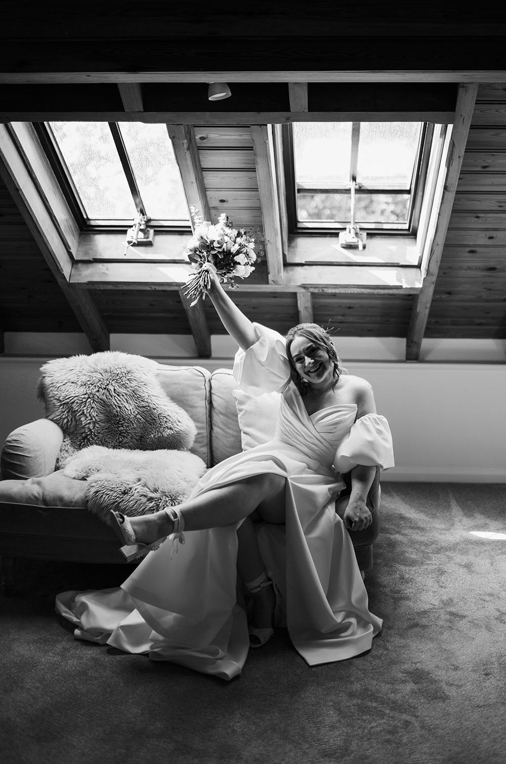Bride in a Satin Faille puff sleeve gown, holding up a bouquet in an attic room with skylights, shown in black and white. 