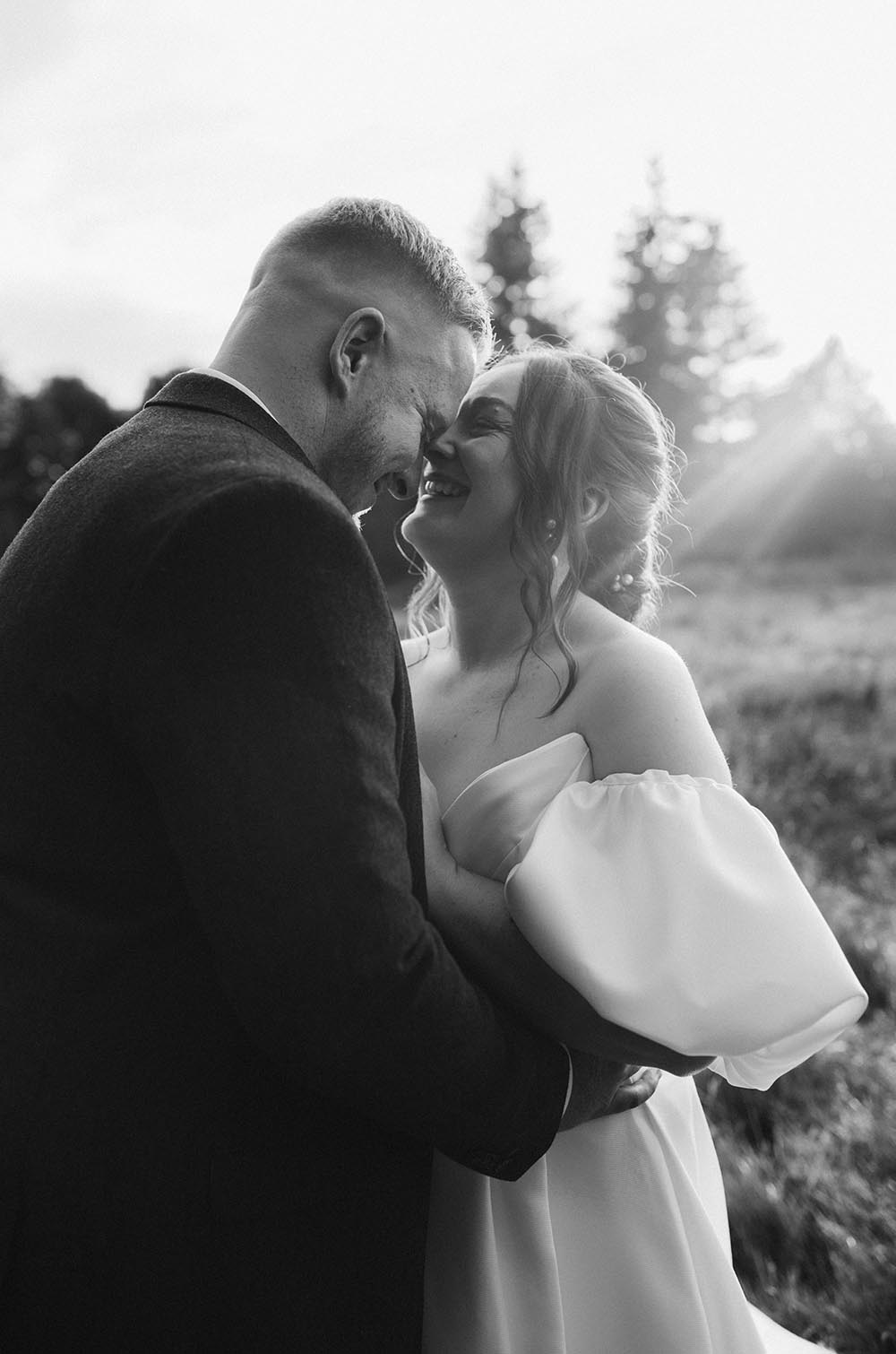 Groom in a black suit embraces his bride in a satin puff sleeve wedding gown, smiling in a meadow in black and white.