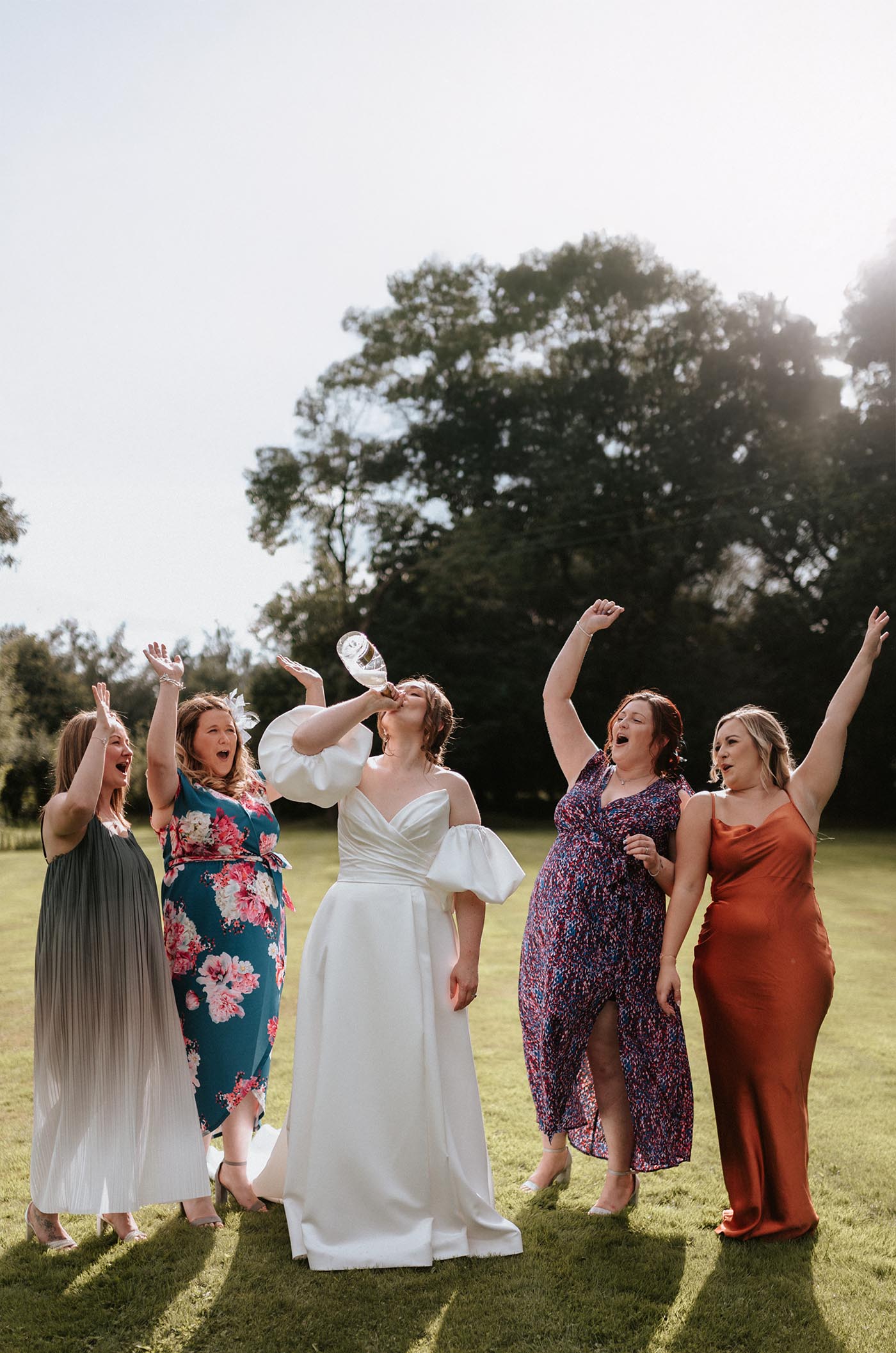 Bride in a strapless satin faille gown with puff sleeves, celebrates alongside bridesmaids cheering in a sunny meadow .