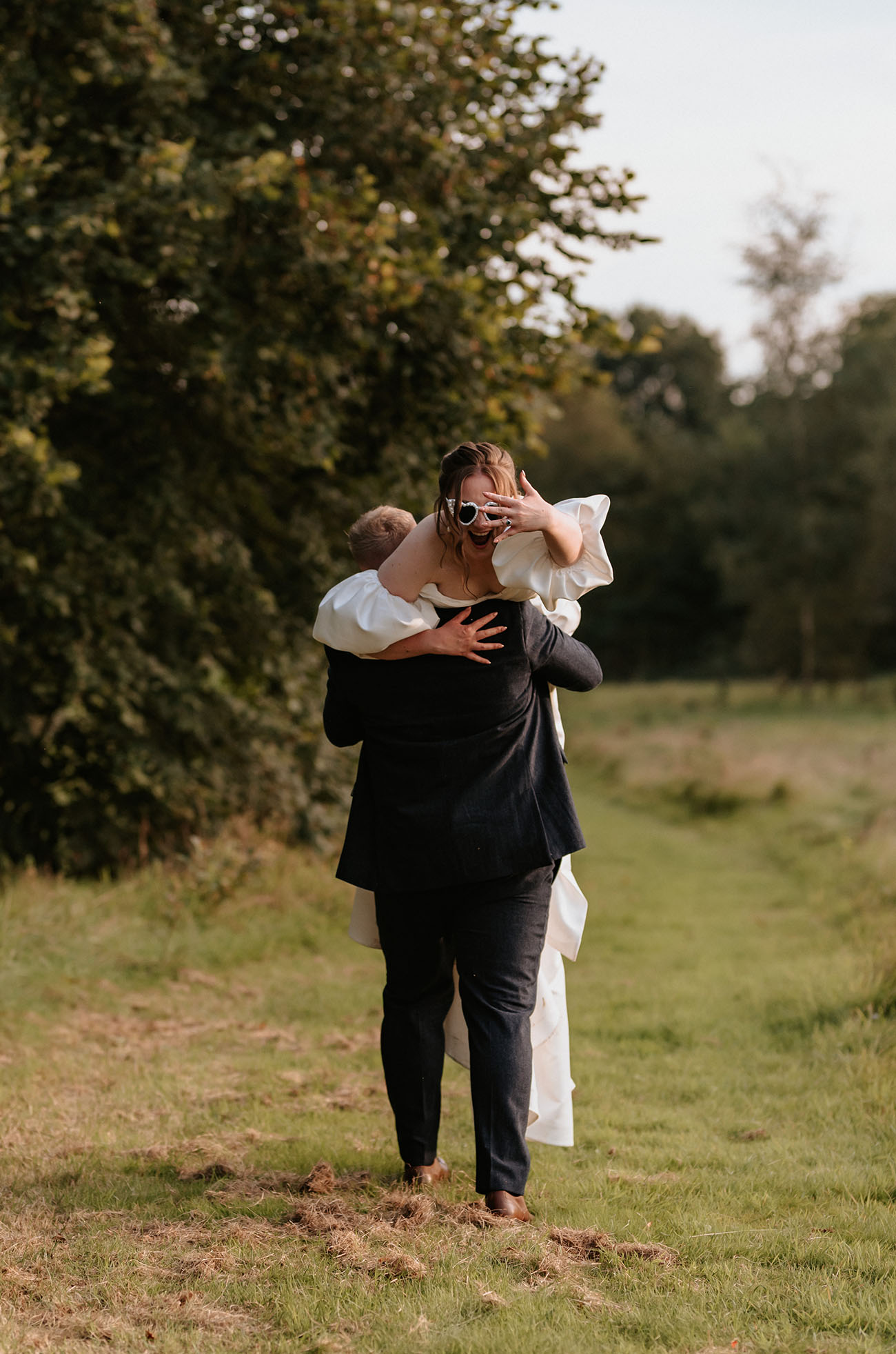 Groom in a black suit picks up his bride in a satin faille gowm with puff sleeves, in a countryside meadow.