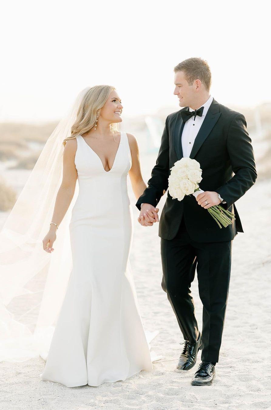 Bride in a fit and flare crepe wedding dress with a V-neckline and veil, smiles holding hands with groom on a sunny beach.