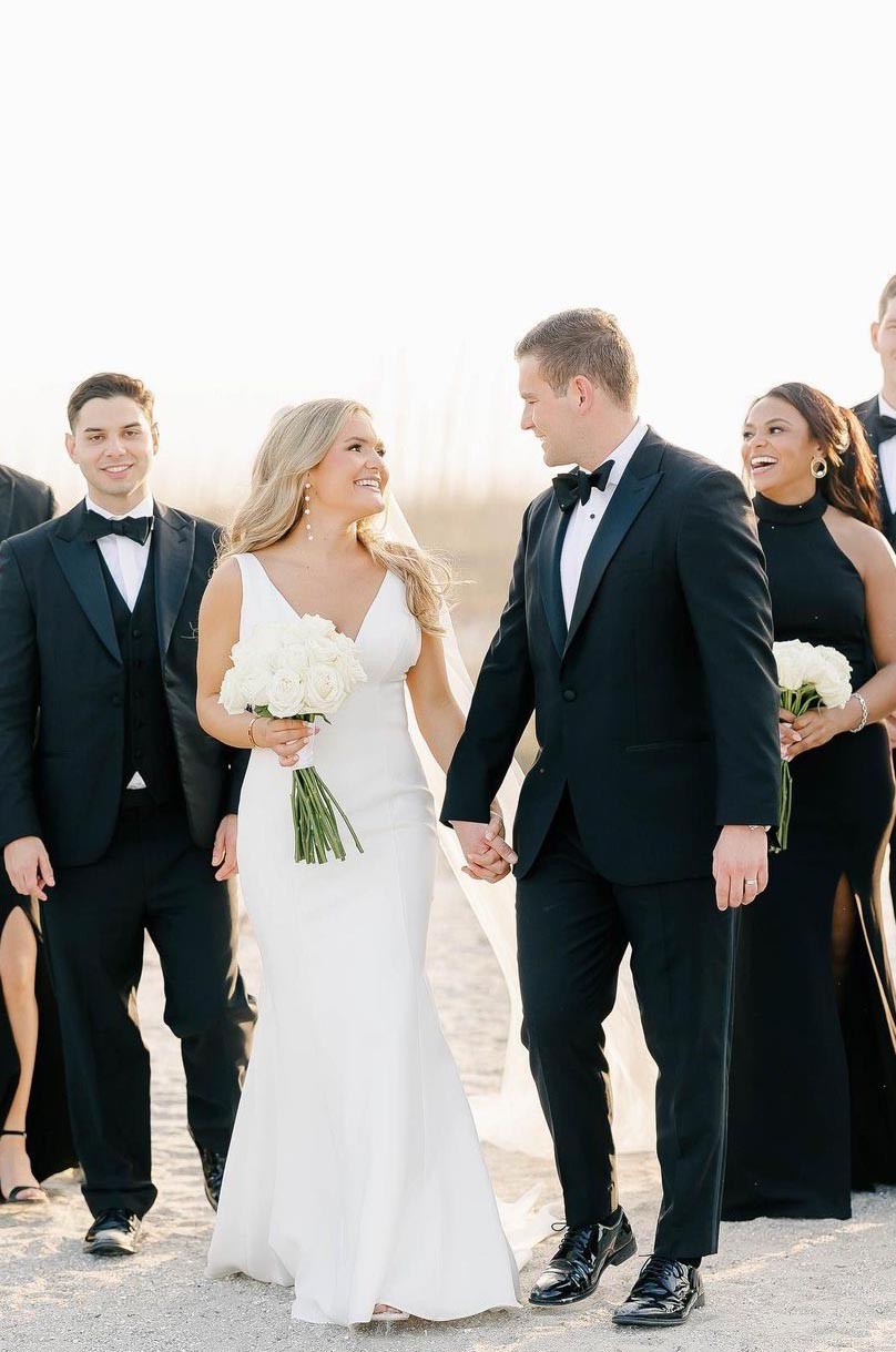 Bride in a sleek V-neck crepe gown, holds hands with groom in a tuxedo and a bouquet in front of bridal party on a beach.