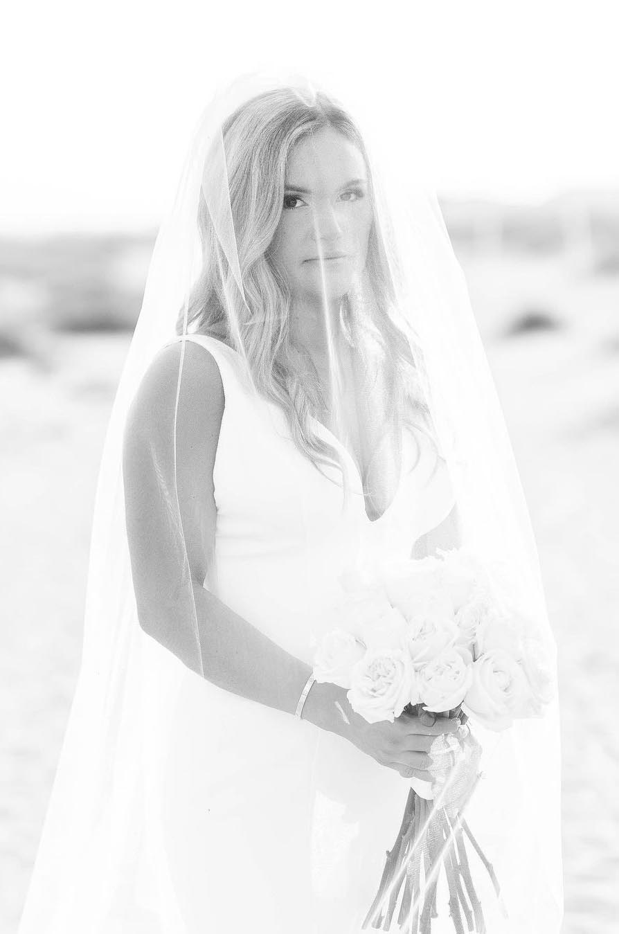 Close-up of bride under a veil, in a plunging V-neck crepe gown, holding a rose bouquet on a beach, in black and white.