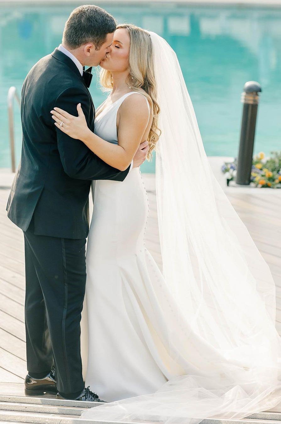 Bride in a fit and flare crepe wedding gown with a plunging V-neckline and a veil, kisses groom by a pool.