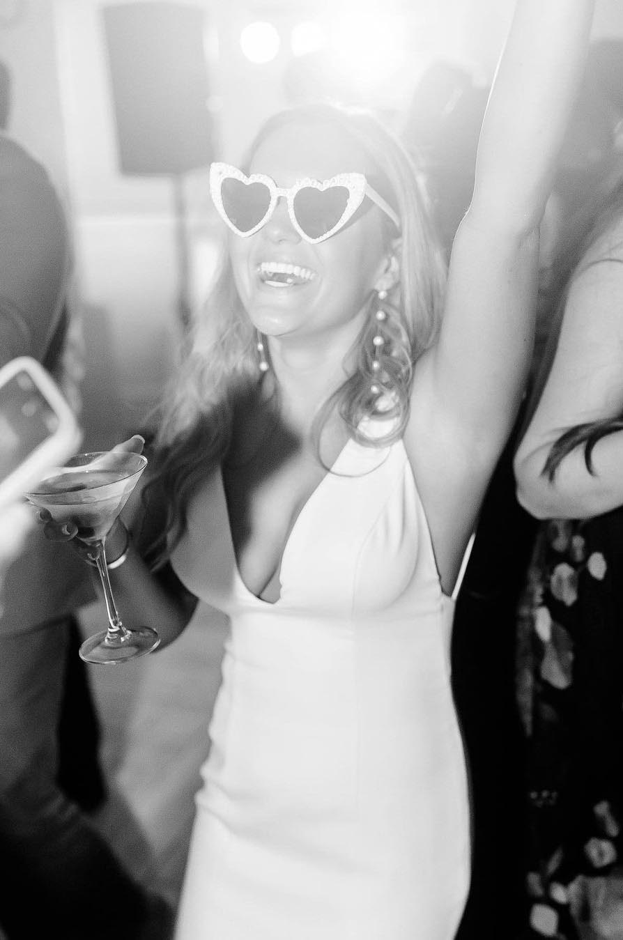 Bride in a fit and flare gown with a deep V-neckline, dances at the reception and is wearing heart-shaped sunglasses.