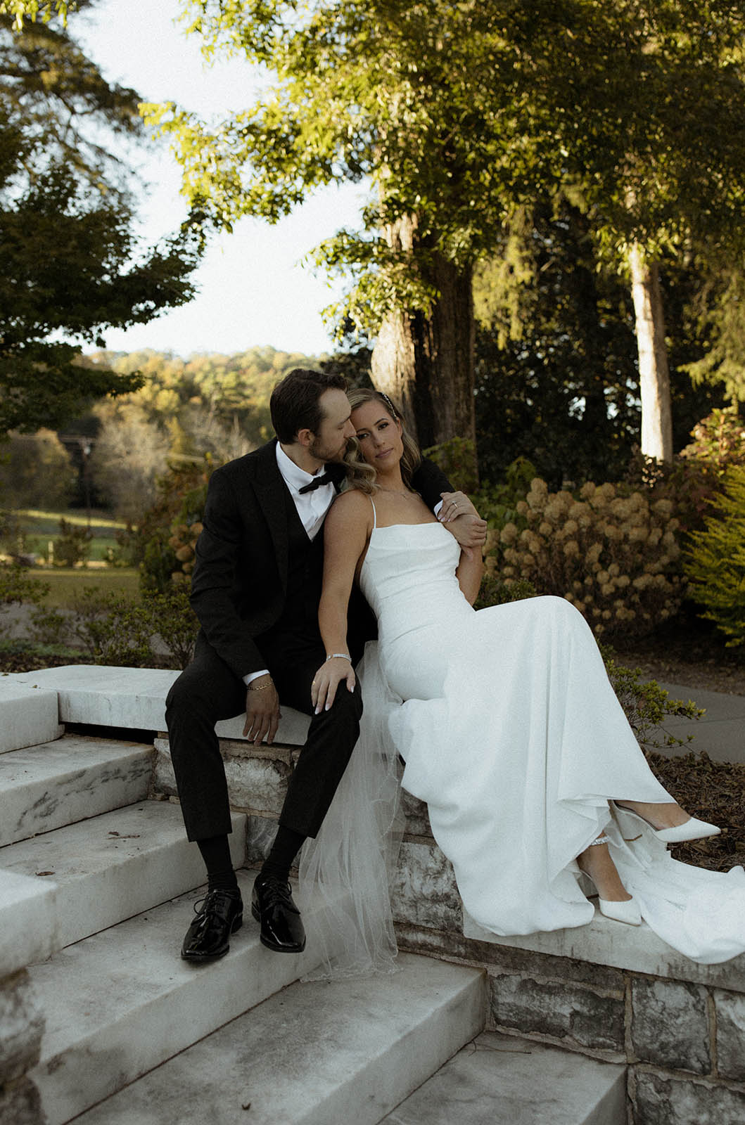 Groom embraces bride in a crepe spaghetti strap wedding dress with a fit and flare skirt, on stone steps outdoors.