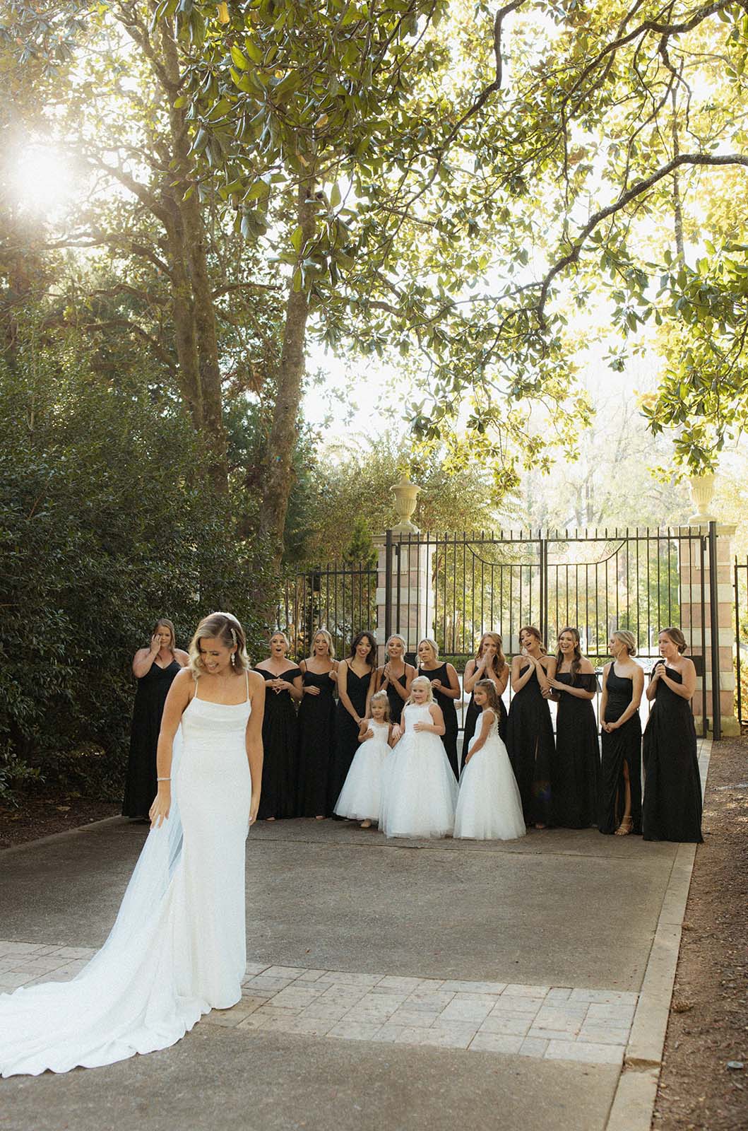 Bride in a spaghetti strap crepe wedding gown poses in front of her bridal party and flower girls at a tall iron gate.