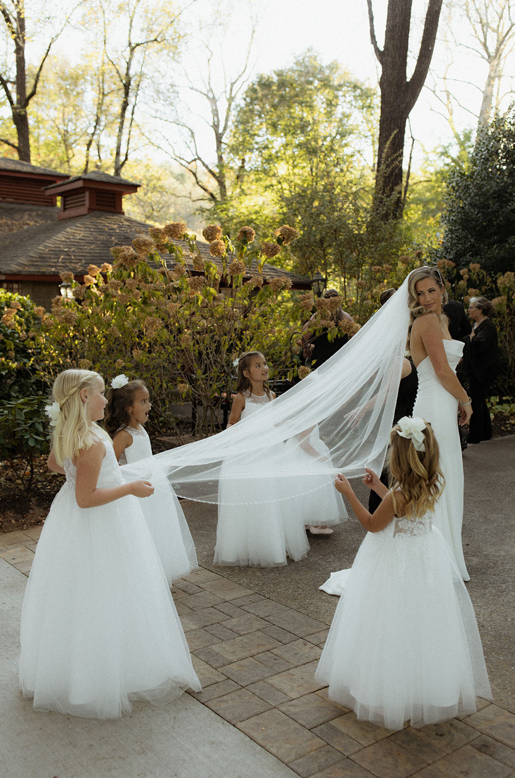 Bride in a scoop neck fit and flare gown with a veil held by flower girls in white dresses, standing outdoors.
