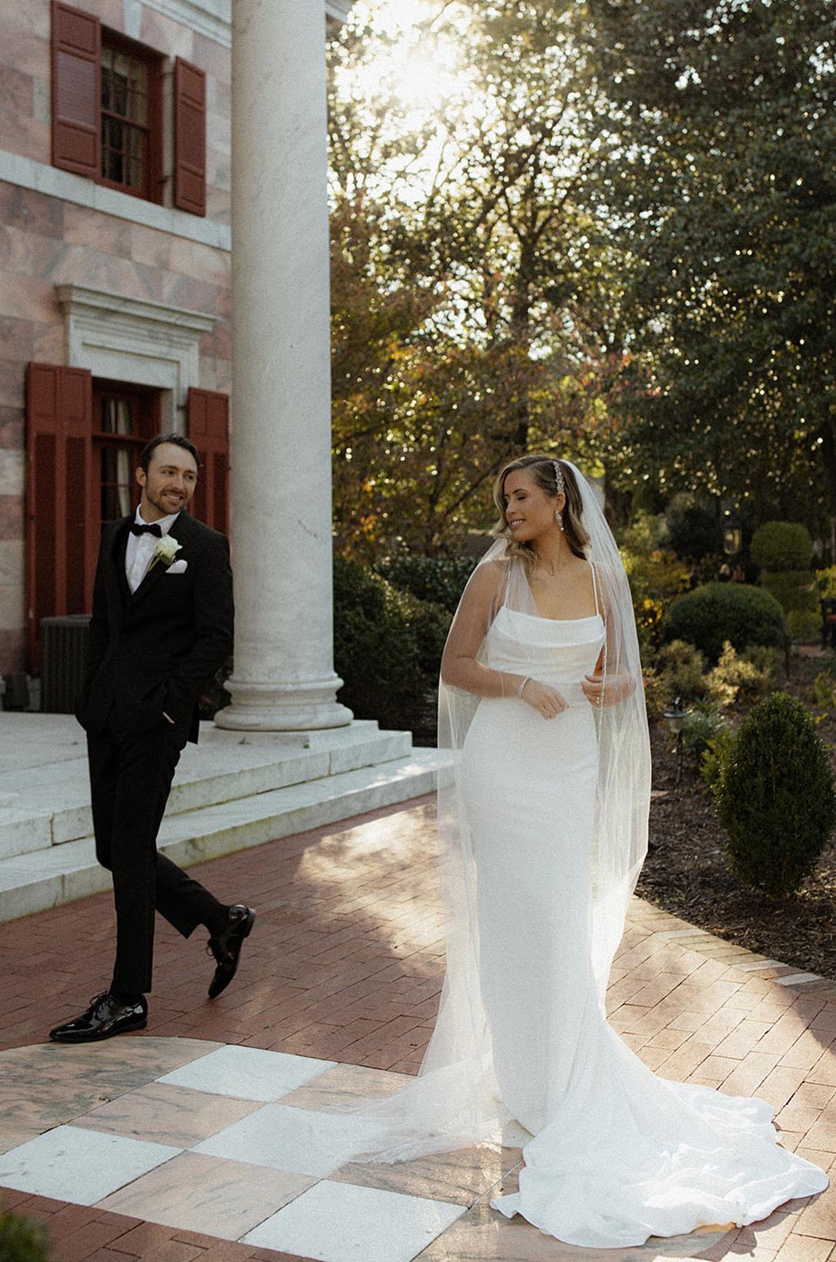 Bride poses in a scoop neck fitted gown with a veil, near groom in a tuxedo outside the venue. 