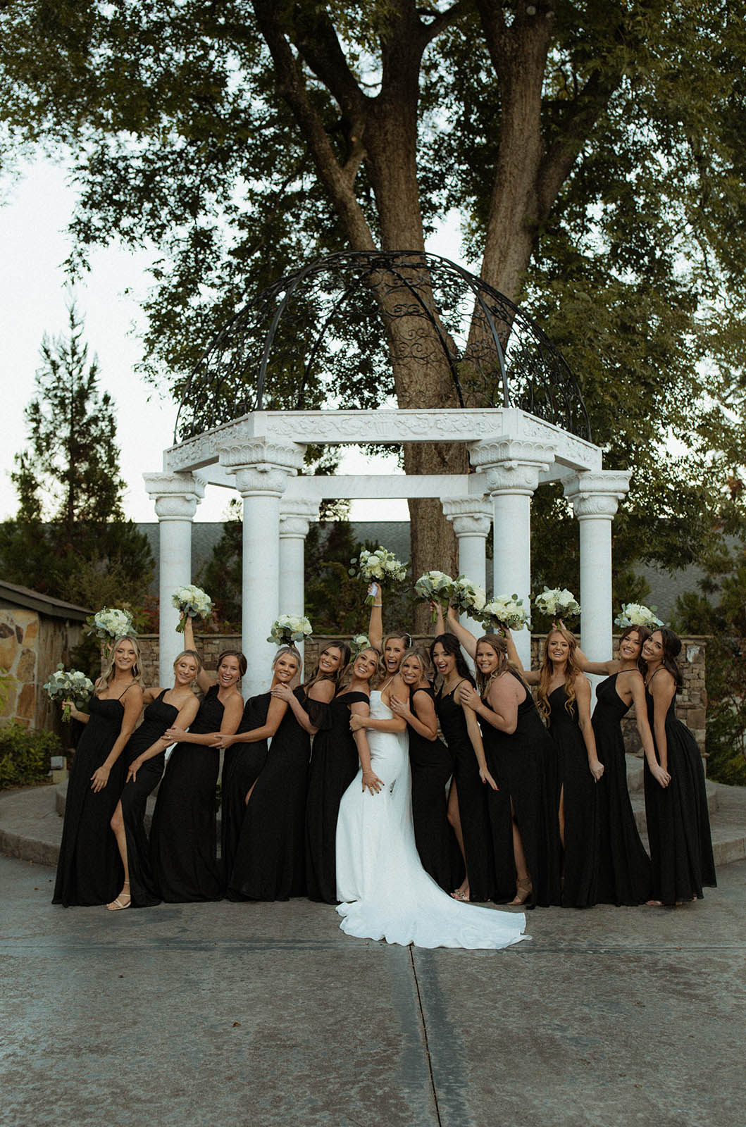 Bride in a fit and flare gown, holds a bouquet up in a line of bridesmaids wearing black dresses at a gazebo with columns.