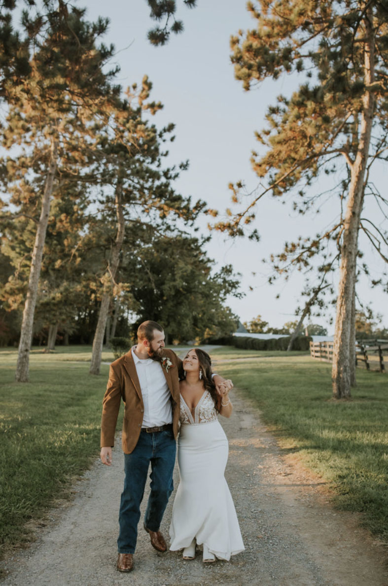 Bride walks alongside groom on a countryside road, in a lace bodice gown with a plunging neckline and a crepe fit and flare skirt.