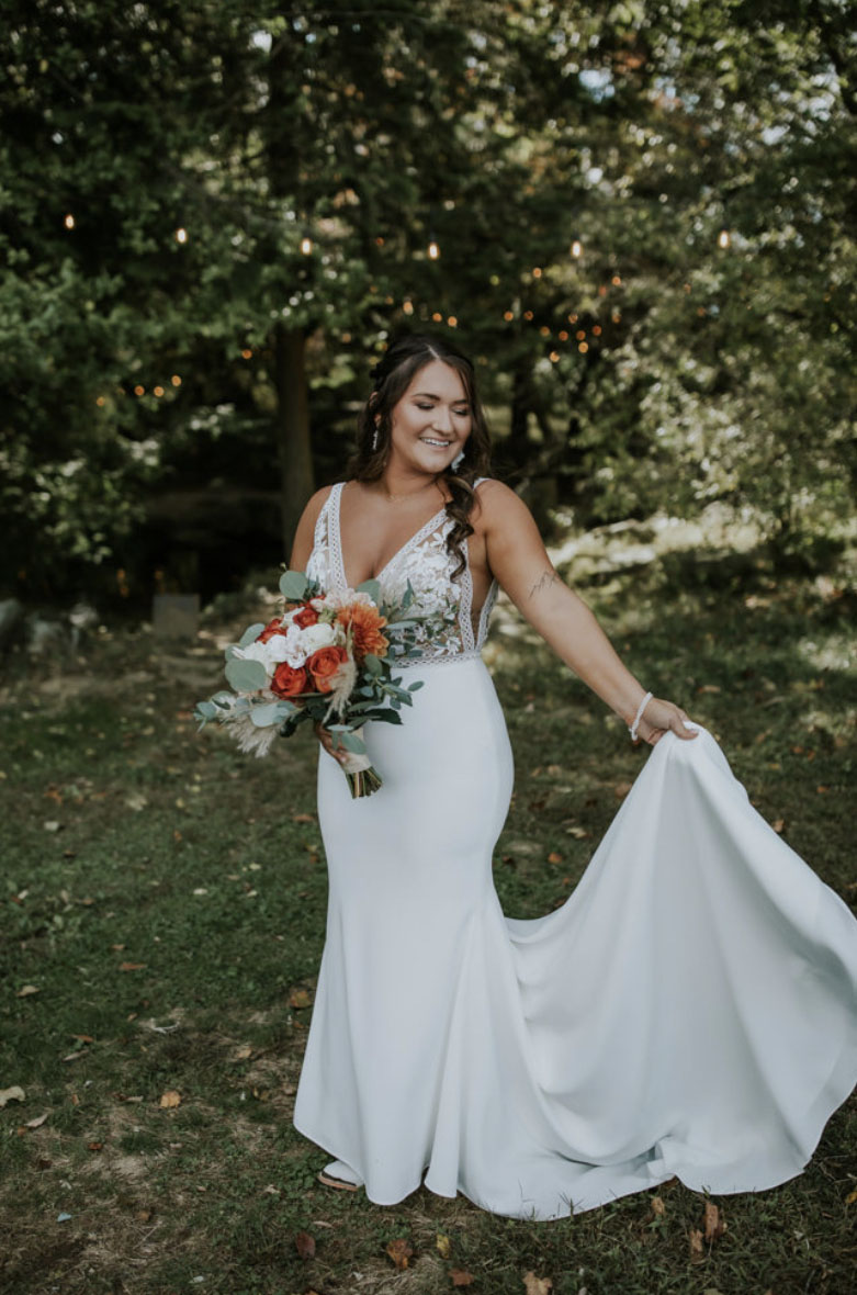 Bride picks up her train in a sleeveless lace bodice wedding dress with a fitted crepe skirt and is holding a bouquet.