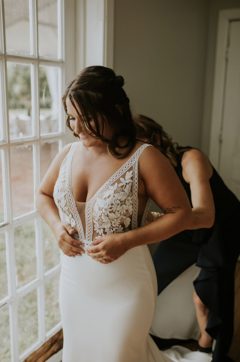 Close-up of smiling bride indoors by a bright window in a deep plunging neck lace gown with a crepe fit and flare skirt.