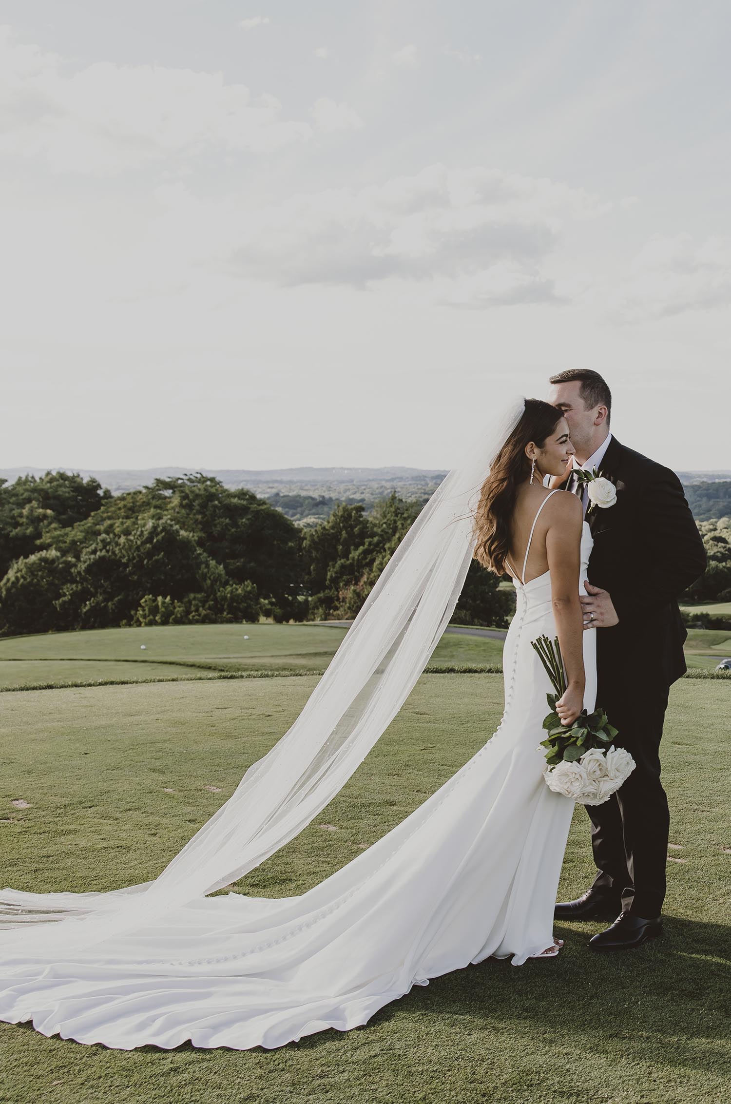 Groom embraces his bride in a fit and flare crepe gown with thin straps, a buttoned back and a veil on a golf course. 