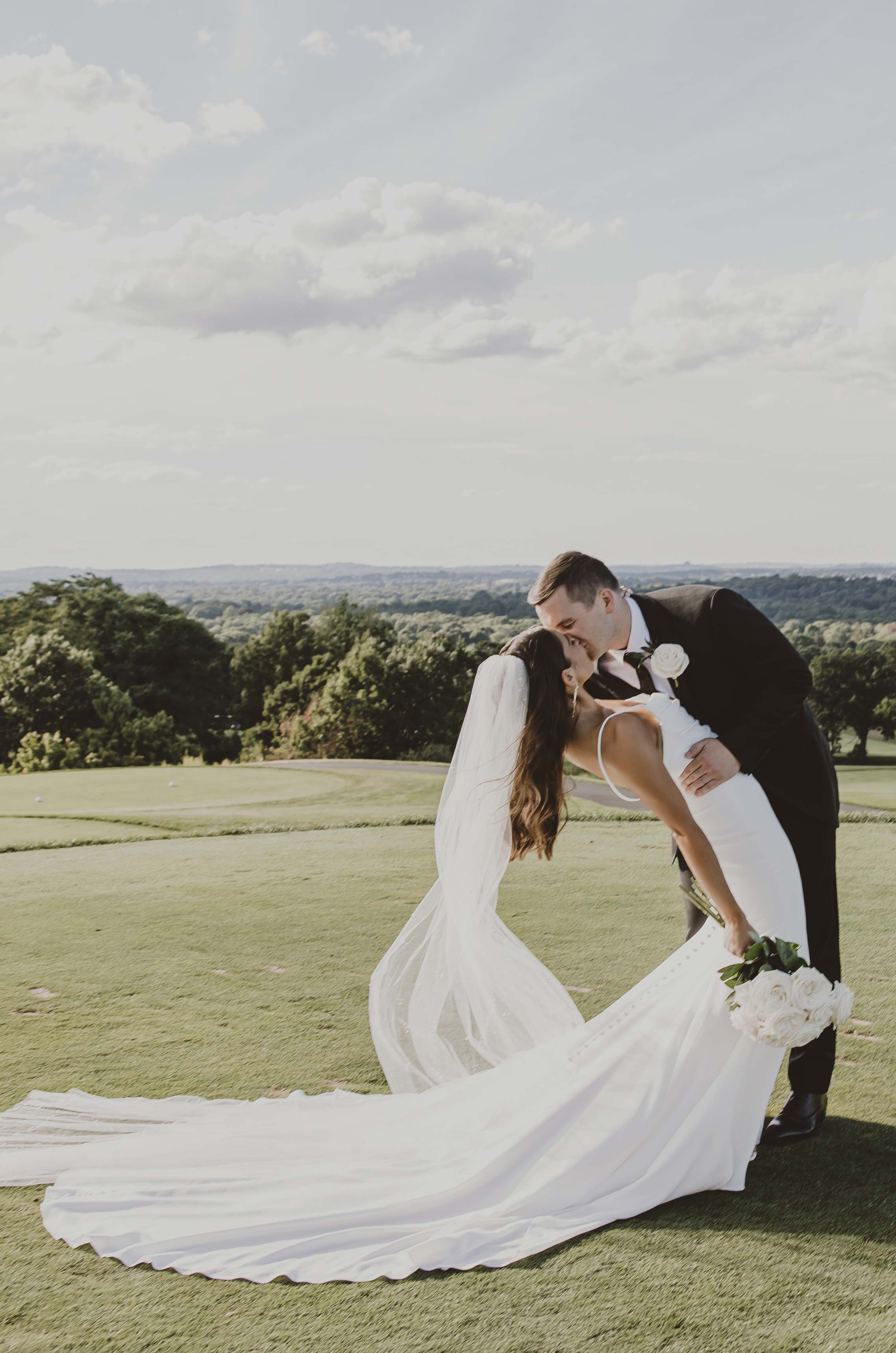Groom in a black suit dips his bride in a sleek fit and flare wedding dress with a veil, holding a bouquet on a hilltop.