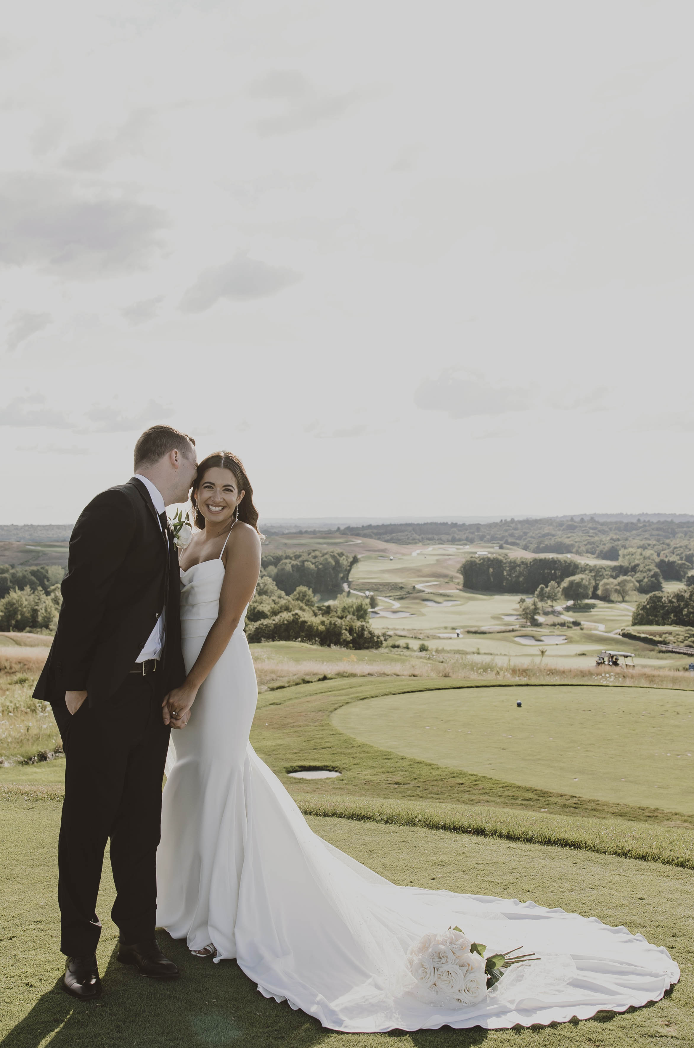Bride in a crepe fitted gown with thin straps and a bouquet at her train poses with groom on an elevated golf course hill.