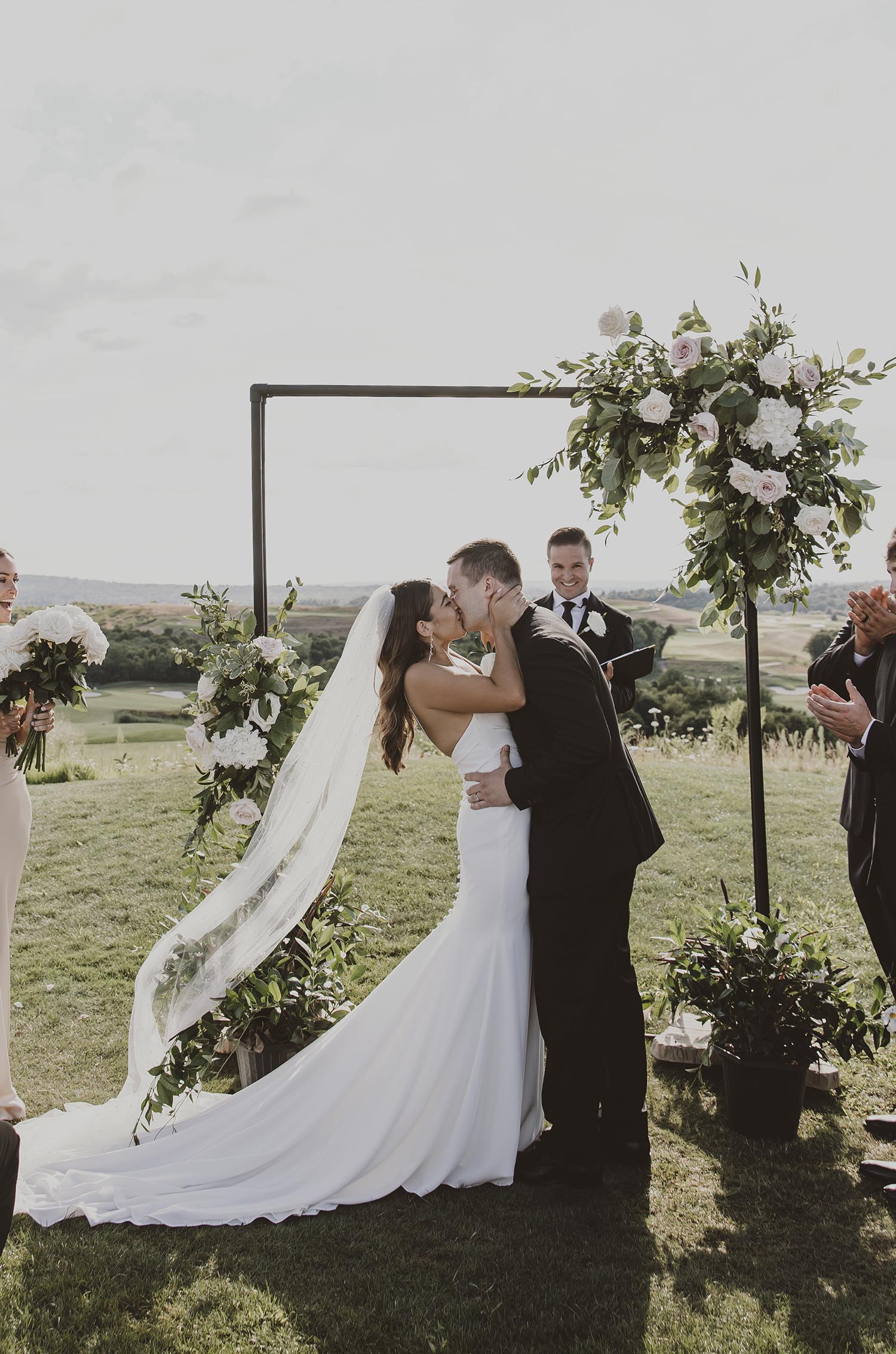 Groom kisses his bride in a crepe fit and flare gown with a flowing train and veil, at altar at outdoor hillside ceremony. 