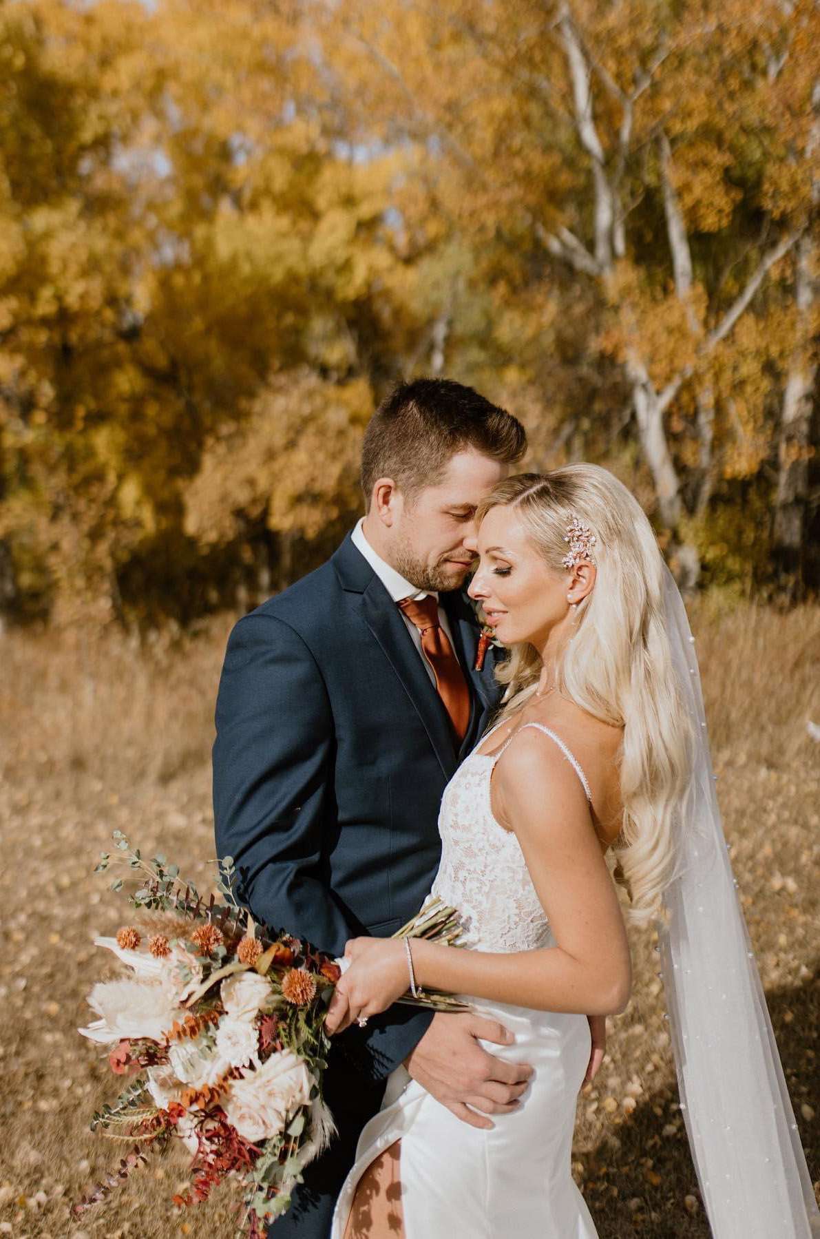 Groom embraces his bride in a lace and satin wedding dress with a slit skirt in a field with fall foliage.