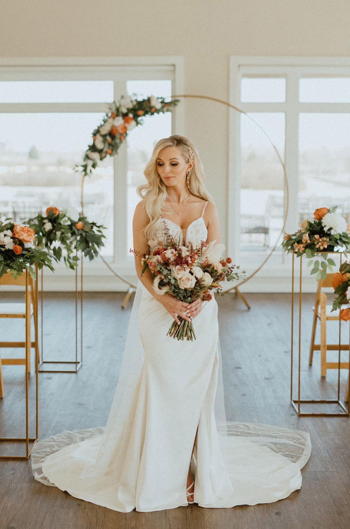 Bride poses in a lace bodice gown with a plunging neck and satin skirt, with a bouquet by large windows and modern decor.