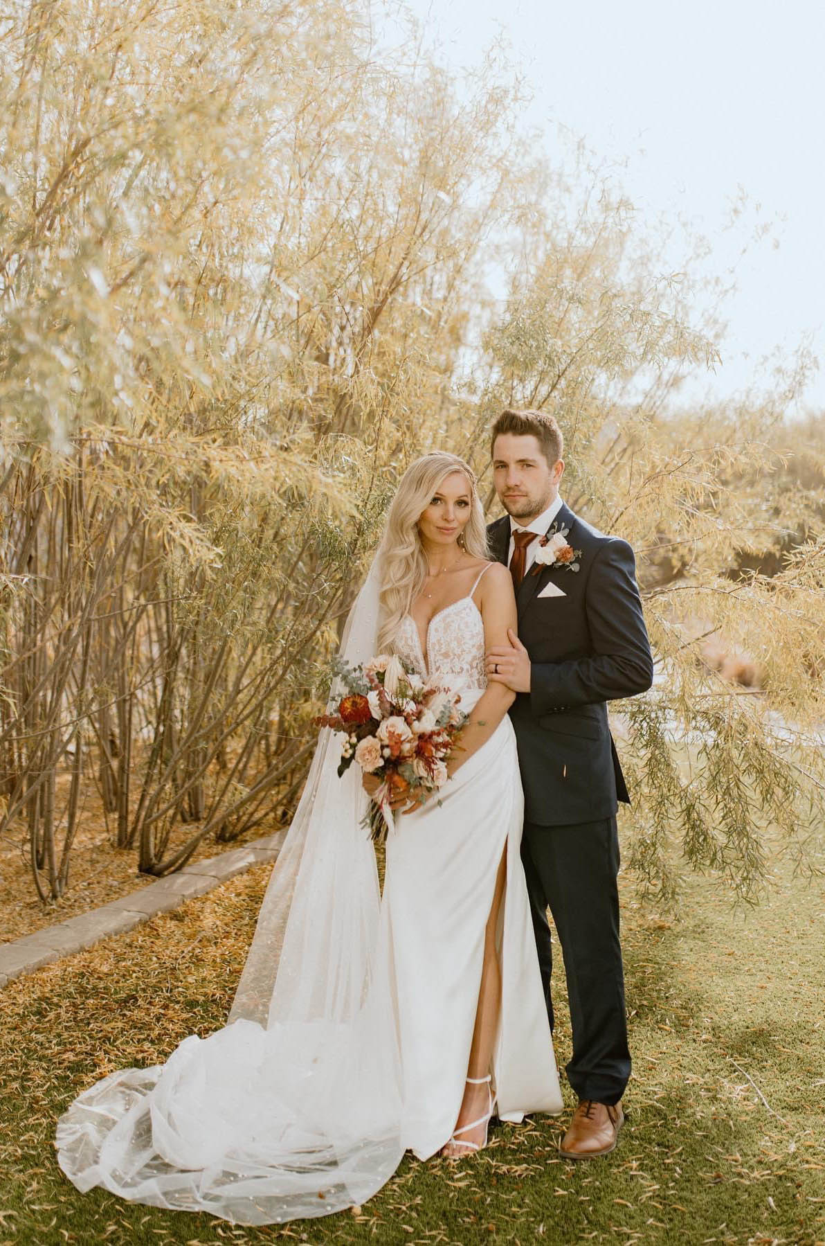 Groom poses next to bride in a plunging neck lace and satin wedding gown with a slit, holding a bouquet by golden willow trees.