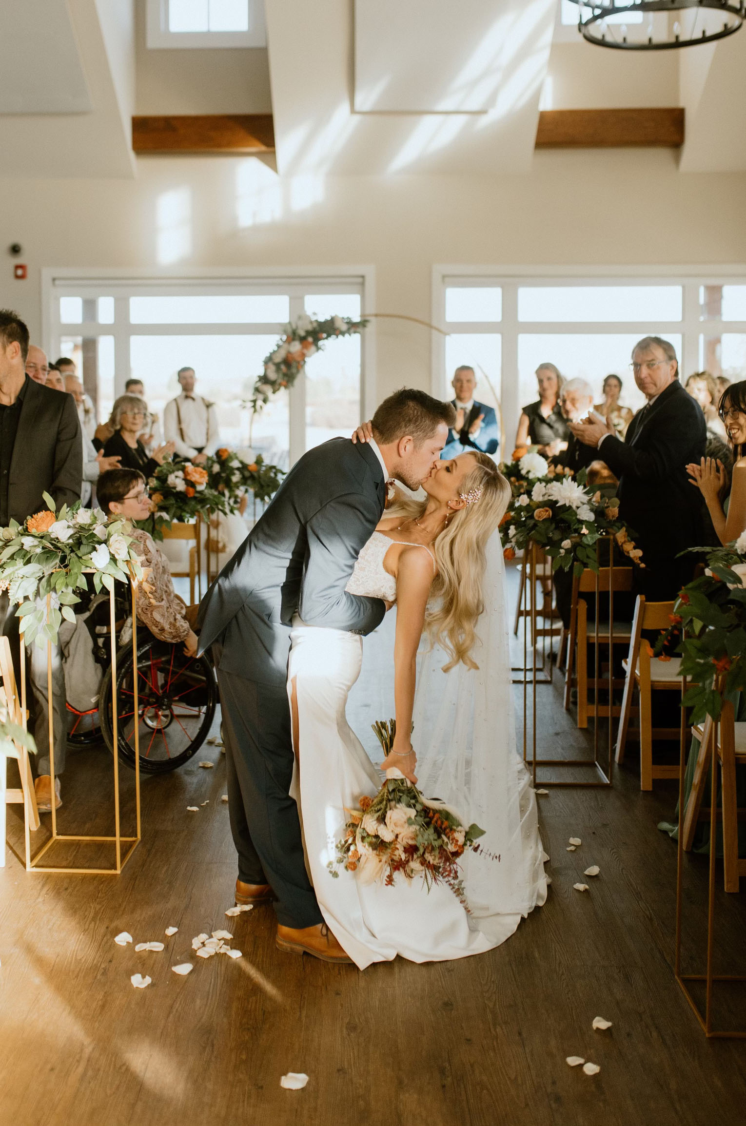  Groom dips his bride in a lace bodice gown with satin slit skirt, thin straps and a veil, holding flowers in the aisle.