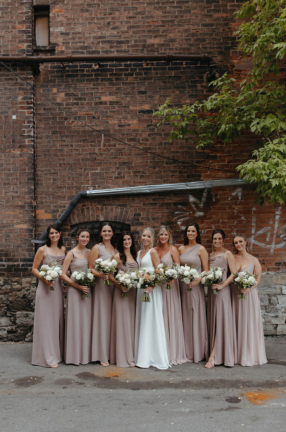 Bride in a sleeveless crepe wedding gown, smiling in a line of bridesmaids with bouquets outside the venue.