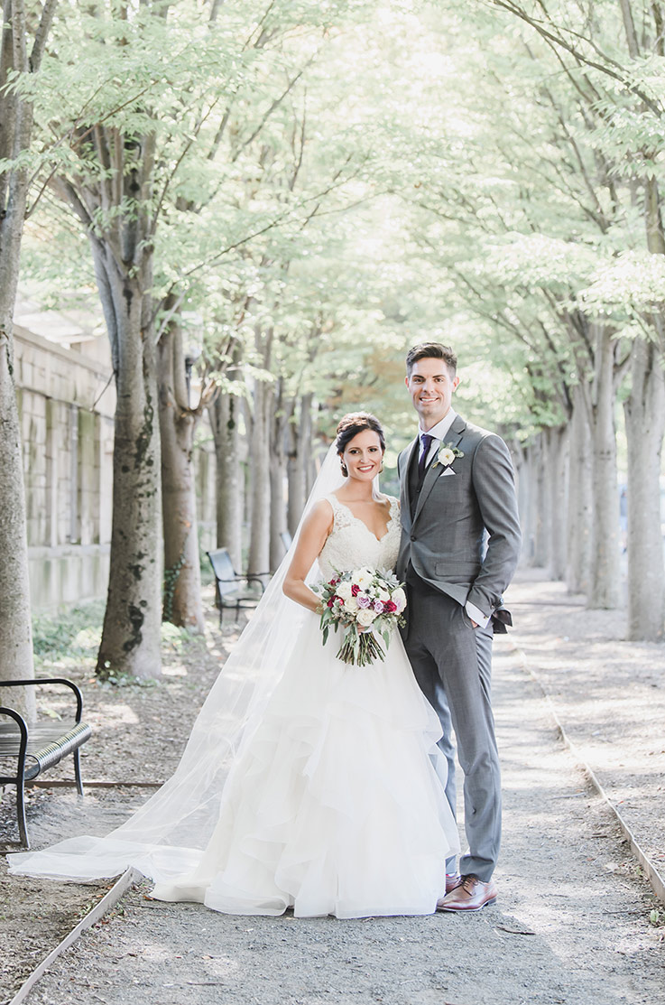 A bride in a lace and tulle dress stands beside a groom in a suit on a path that is between two rows of trees.