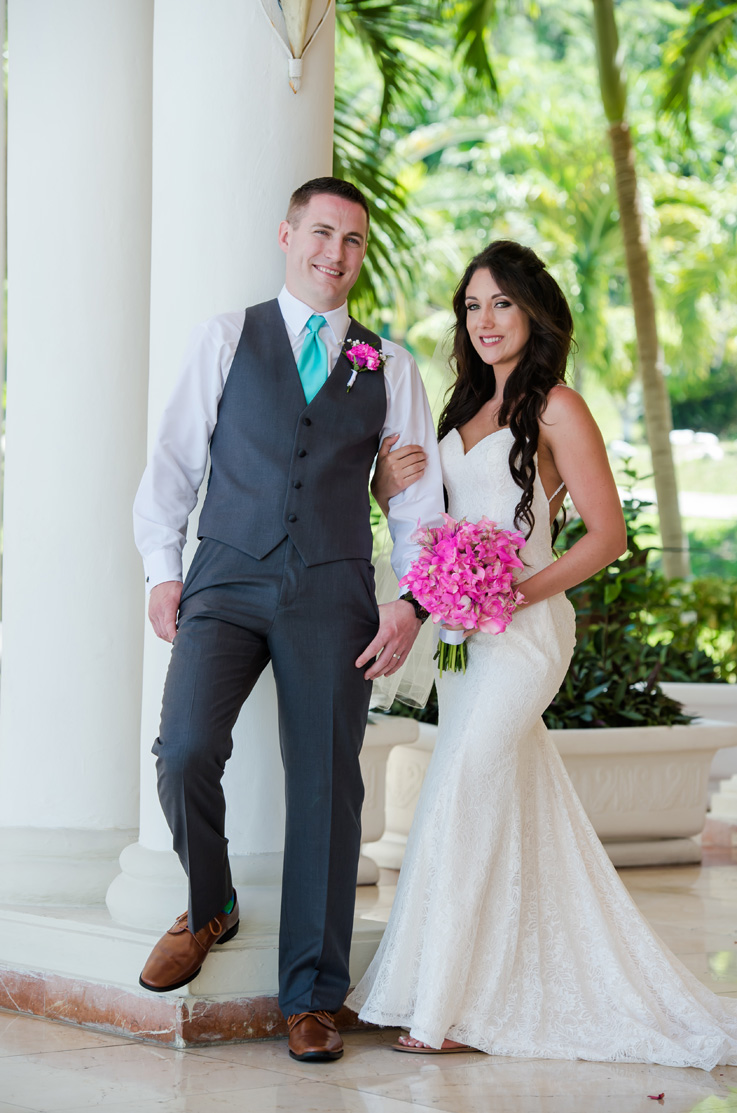 A bride in a lace wedding dress holds a bouquet of flowers in one hand and a groom's arm in the other, while standing outside in front of pillars and tropical trees.