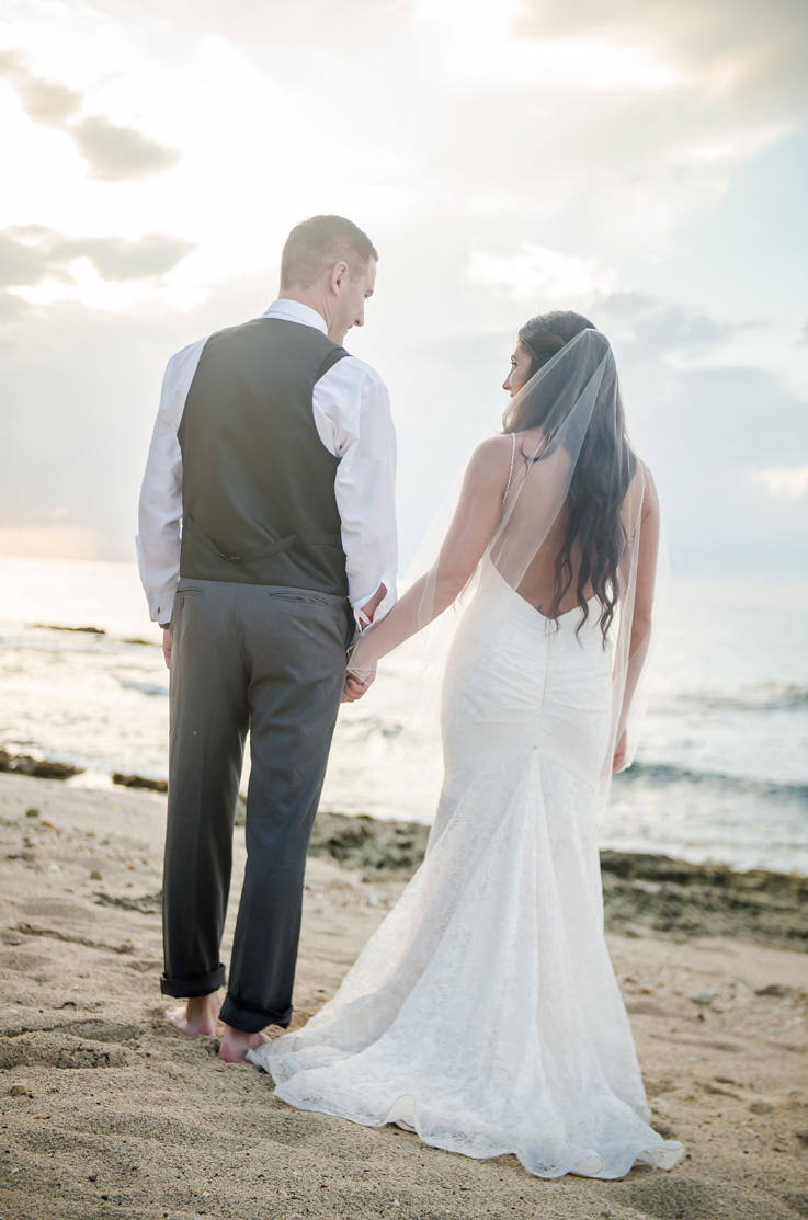 A bride in a lace wedding dress and veil, and a groom in a suit vest, stand on a beach facing the waves and looking at each other.