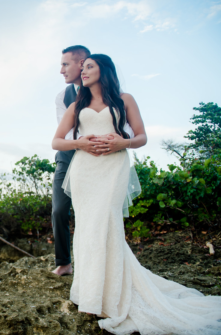 A groom in a suit vest wraps his arms around a bride in a lace wedding dress outdoors, while they look off to the side.