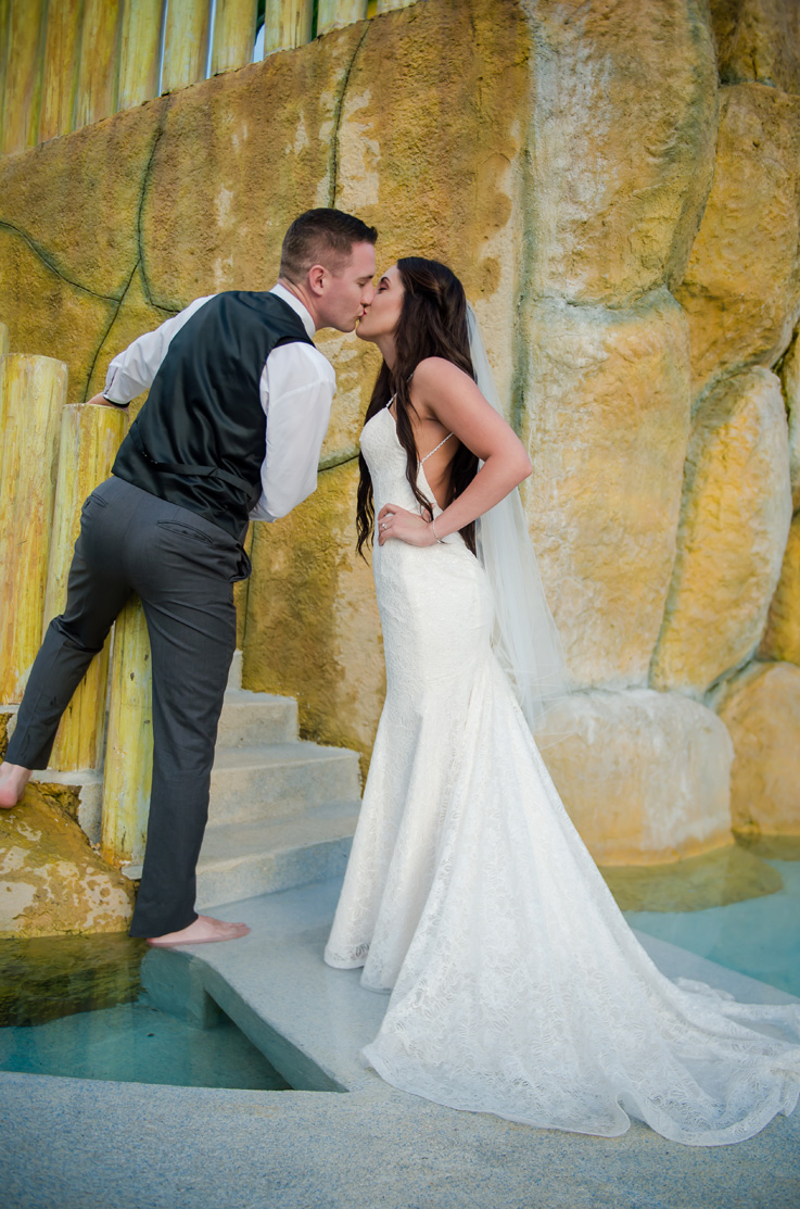 A bride in a lace wedding dress and a groom in a suit vest kiss on a path outdoors.
