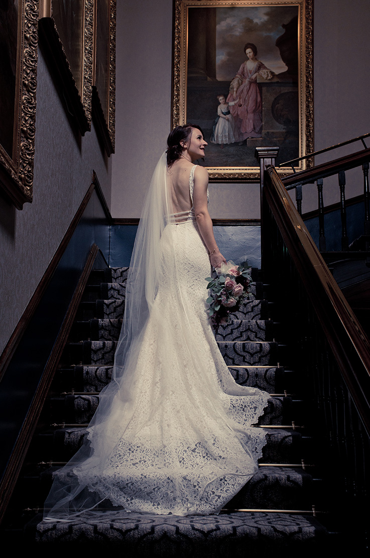 A bride in a lace wedding dress and veil stands on a staircase holding a bouquet of flowers, surrounded by large portraits.
