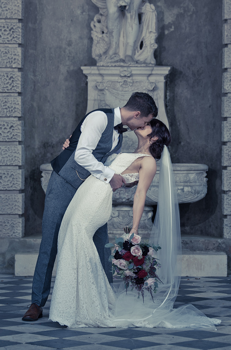 A groom in a suit vest dips and kisses a bride in a lace wedding dress in front of a sculpture.