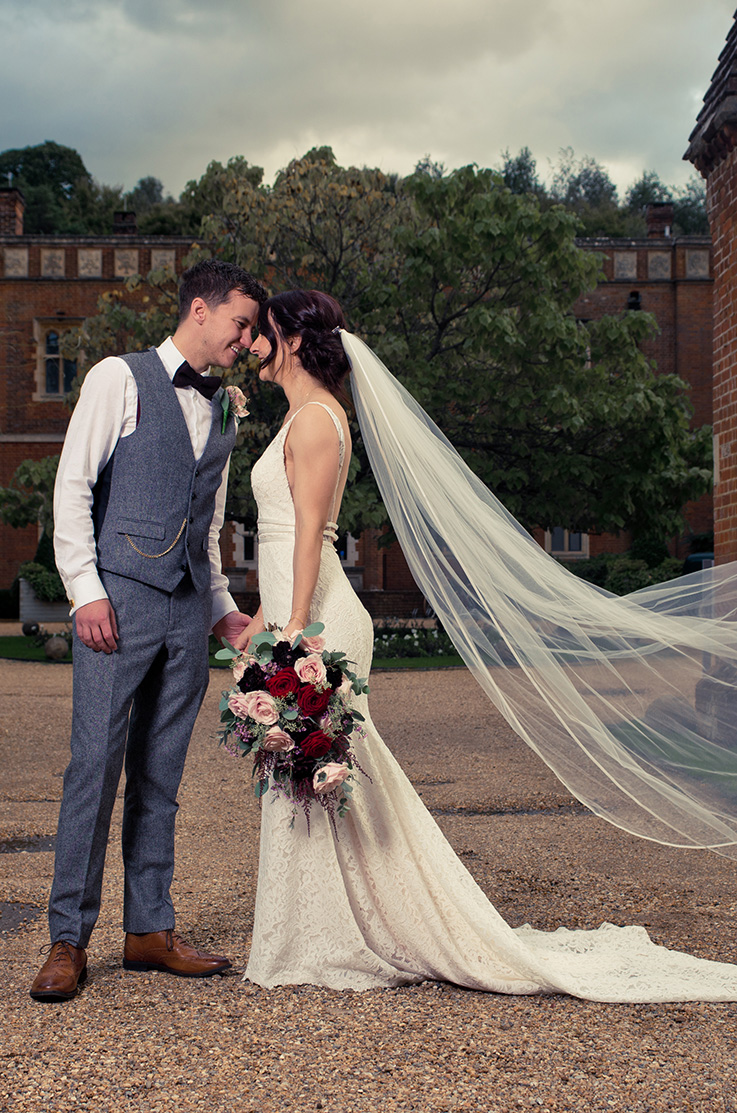 A bride in a fitted lace wedding dress and veil, and a groom in a suit vest and bow tie stand outside in a courtyard while looking at each other and smiling.