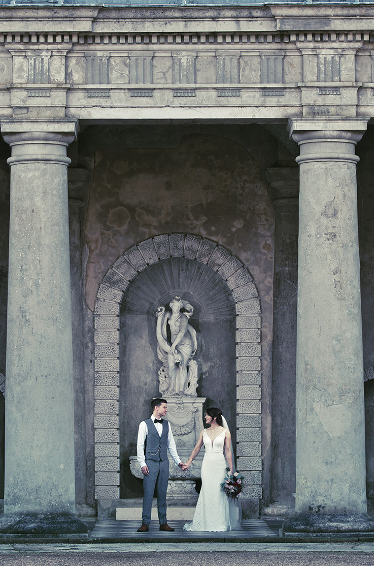 A bride in a fitted lace wedding dress and a groom in a suit vest and bow tie stand in front of a sculpture and large stone pillars while looking at each other and holding hands.