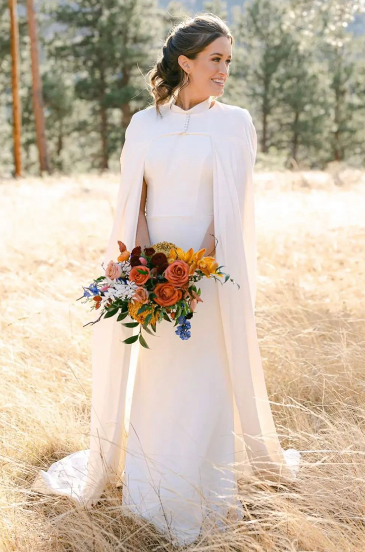 A bride in a fitted wedding dress and a long cape holds a bouquet in both hands, while looking off to the side and smiling, outside in front of a forest.