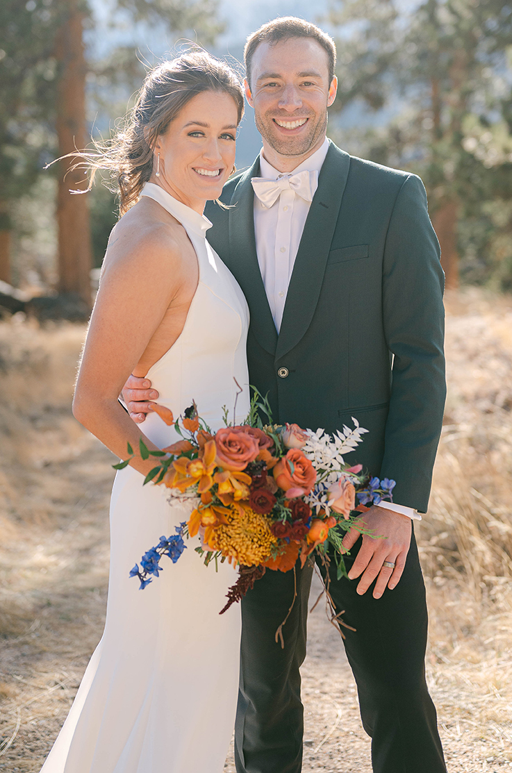 A bride in a fitted wedding dress holds a bouquet of flowers and stands by a groom in a suit, outside in front of a forest.
