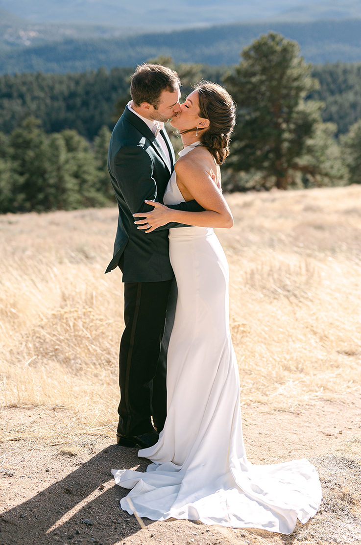 A bride in a fitted wedding dress and a groom in a suit hold each other and kiss in a field of tall yellow grass, with a forest in the background.