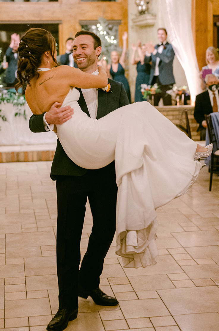 A groom in a suit sweeps a bride in a fitted wedding dress off her feet and smiles in an event hall, while the wedding party cheers in the background.
