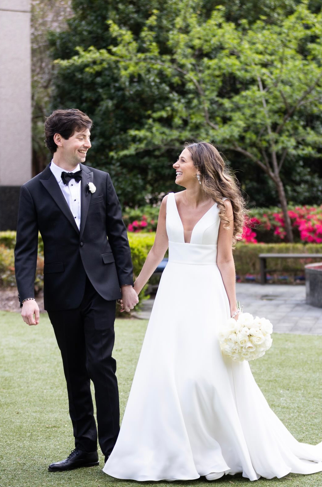 A bride in a sleeveless A-line wedding dress and a groom in a suit and bow tie look at each other while holding hands and smiling, outside in a garden.