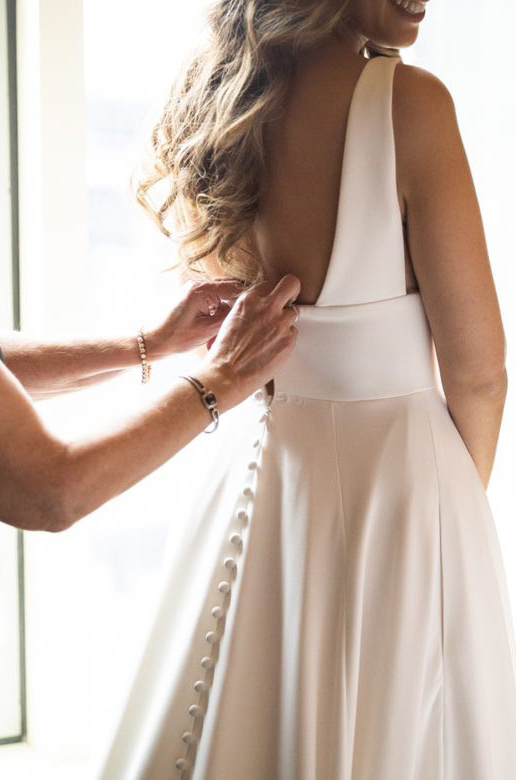 A bride in an A-line wedding dress with buttons down the back gets help closing the back of the dress from a friend.