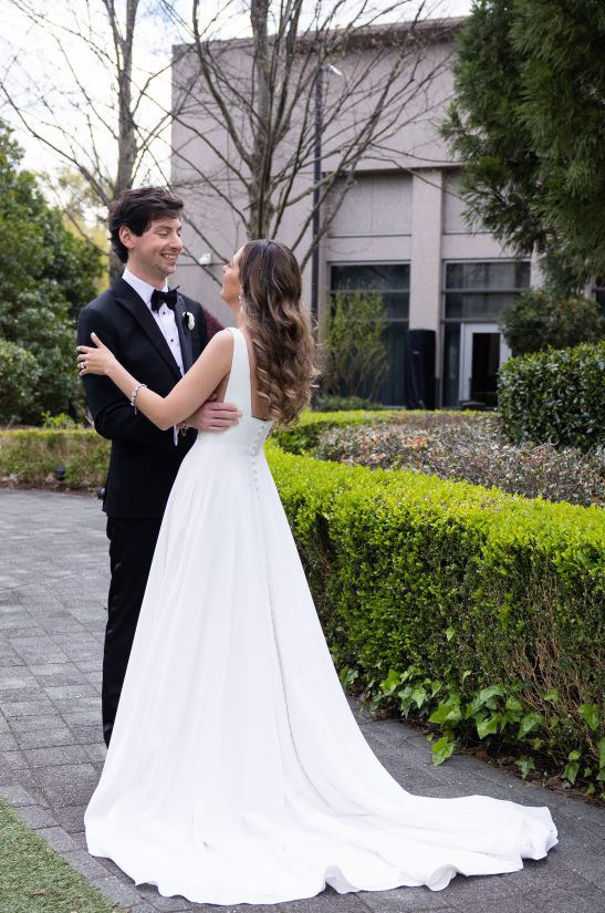 A bride in a sleeveless A-line wedding dress and a groom in a suit and bow tie look into each others eyes and hold each other while standing outside on a path, in front of a hedge.