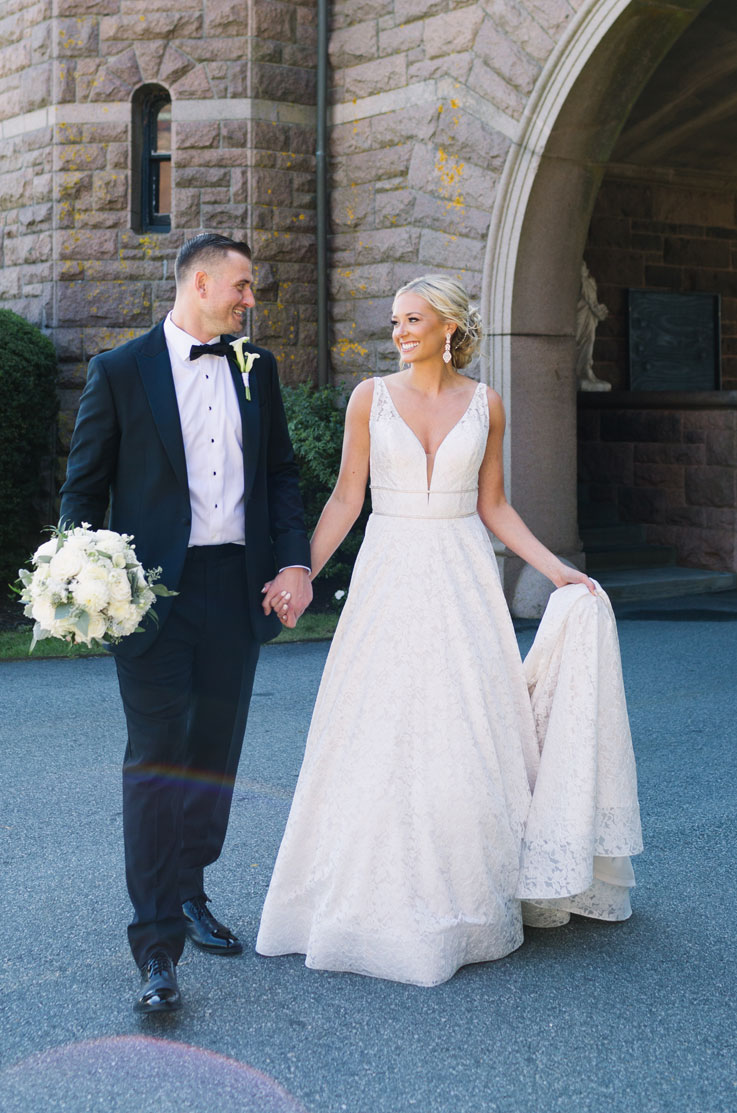 A bride in a lace A-line wedding dress and a groom in a suit stand by each other, holding hands and smiling, outside on a paved road in front of a stone building.
