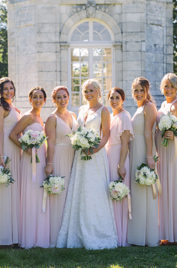 A bride in a lace wedding dress stands in the middle of six bridesmaids, all holding bouquets, outside in front of a stone building.