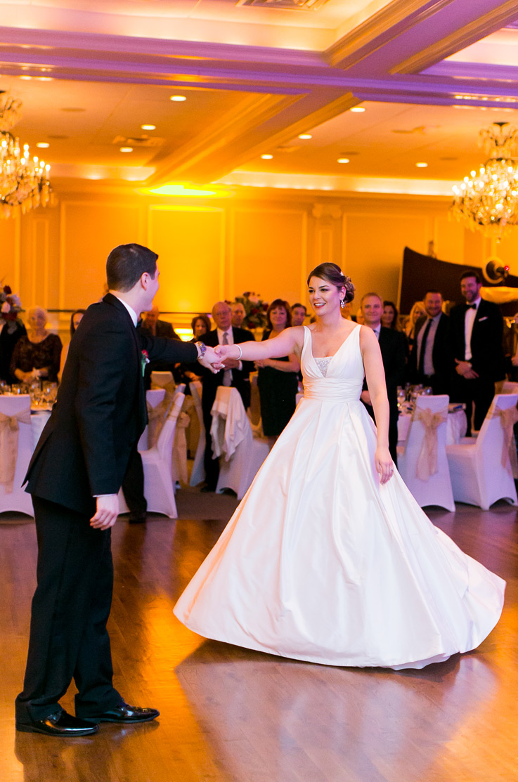 A bride in a V-neckline ball gown wedding dress dances with a groom in a suit on the dance floor, with wedding guests, tables, and chairs in the background.