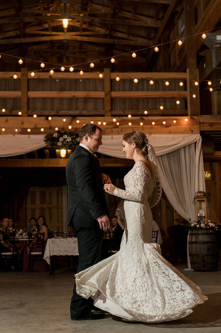 A bride in a bustled long sleeve lace wedding dress and a groom in a suit dance together in a farm house venue while wedding guests watch in the background.